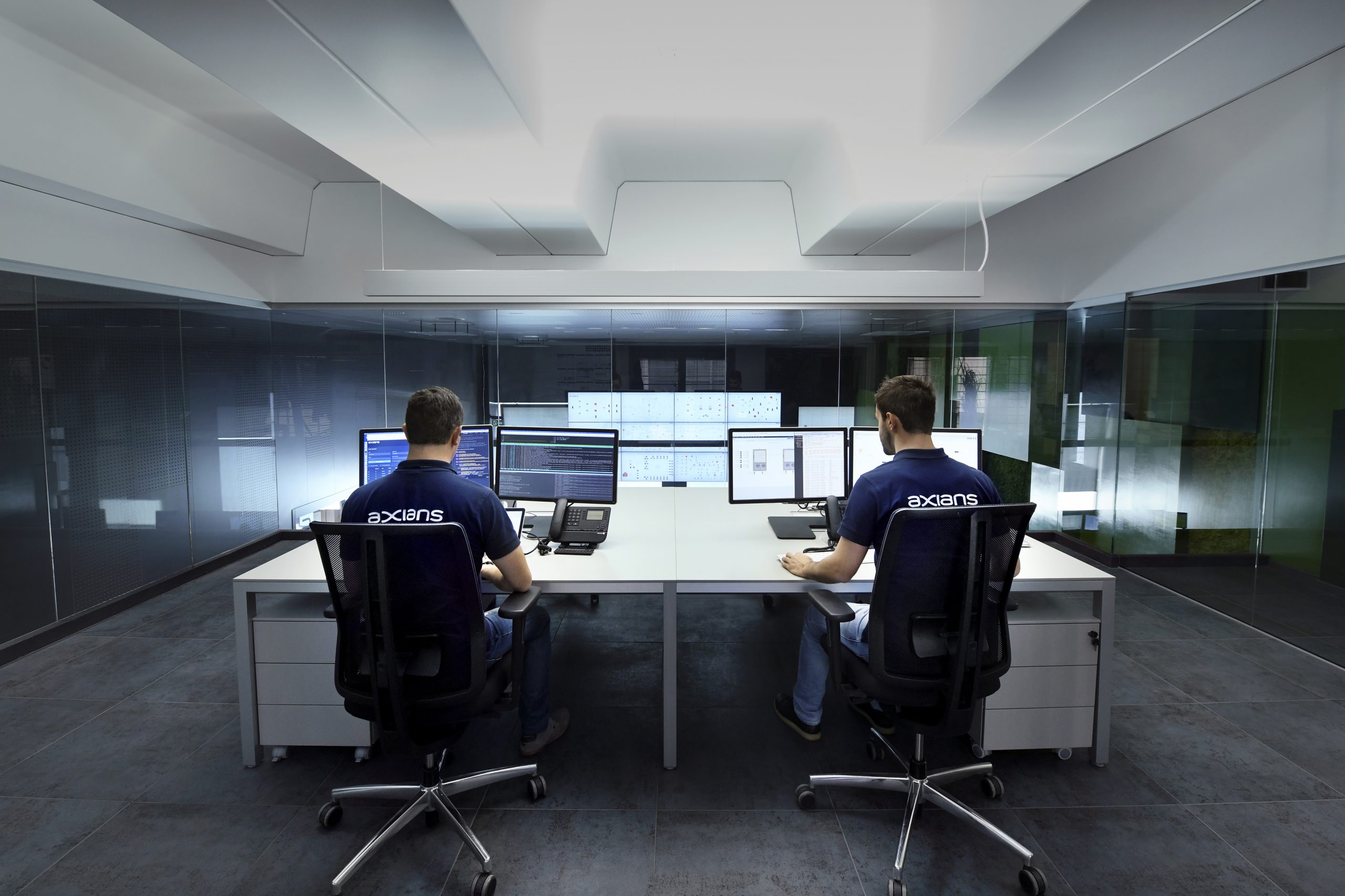 Two Axians cybersecurity analysts monitor live systems and analyze code inside a glass-walled Security Operations Center, supporting real-time threat detection and compliance.