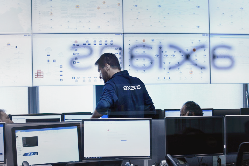 Technician wearing an Axians shirt overseeing network monitoring systems in a control room with multiple computers and a large wall of screens.