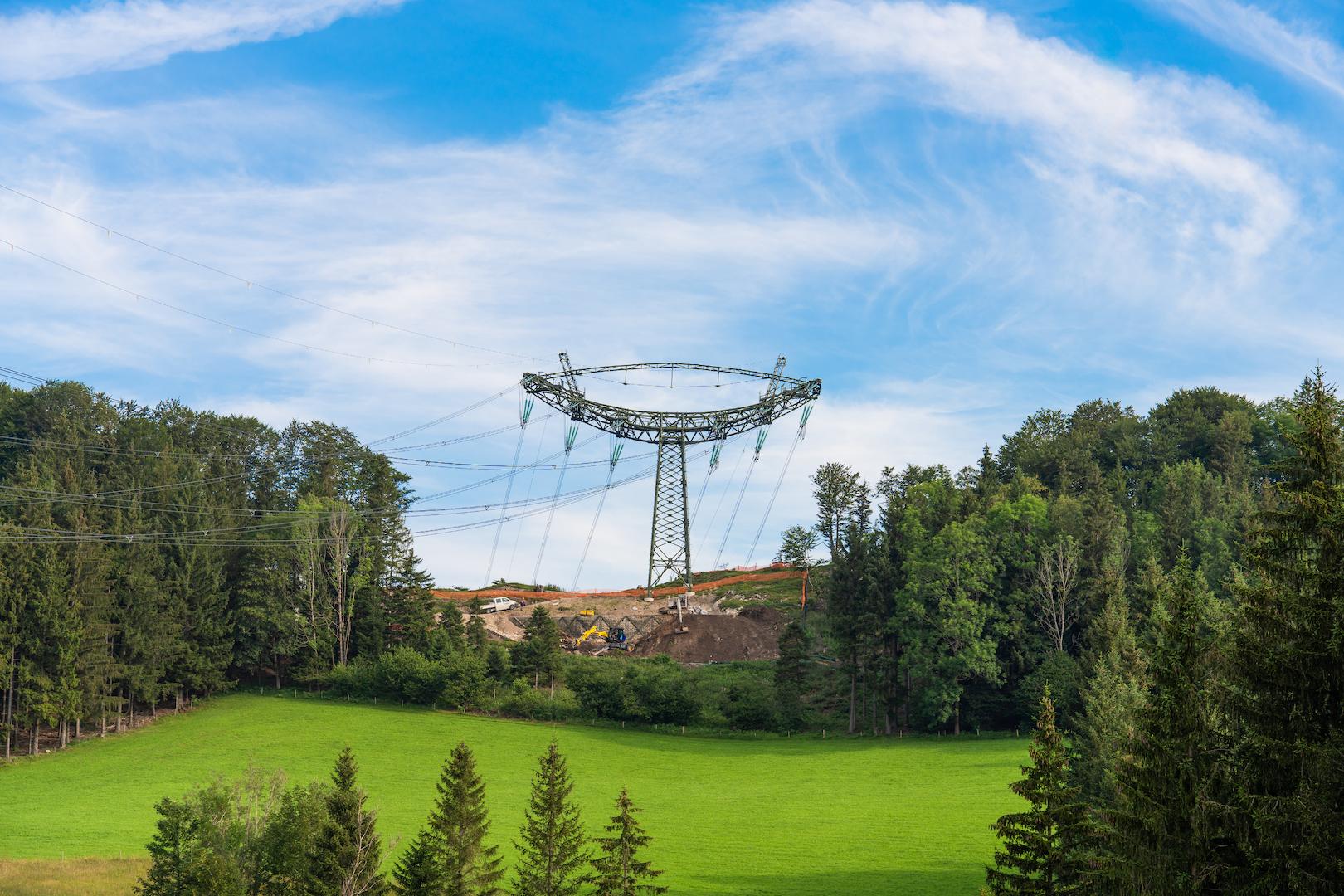 High-voltage overhead transmission line tower installed across green fields and forested hills in an alpine landscape